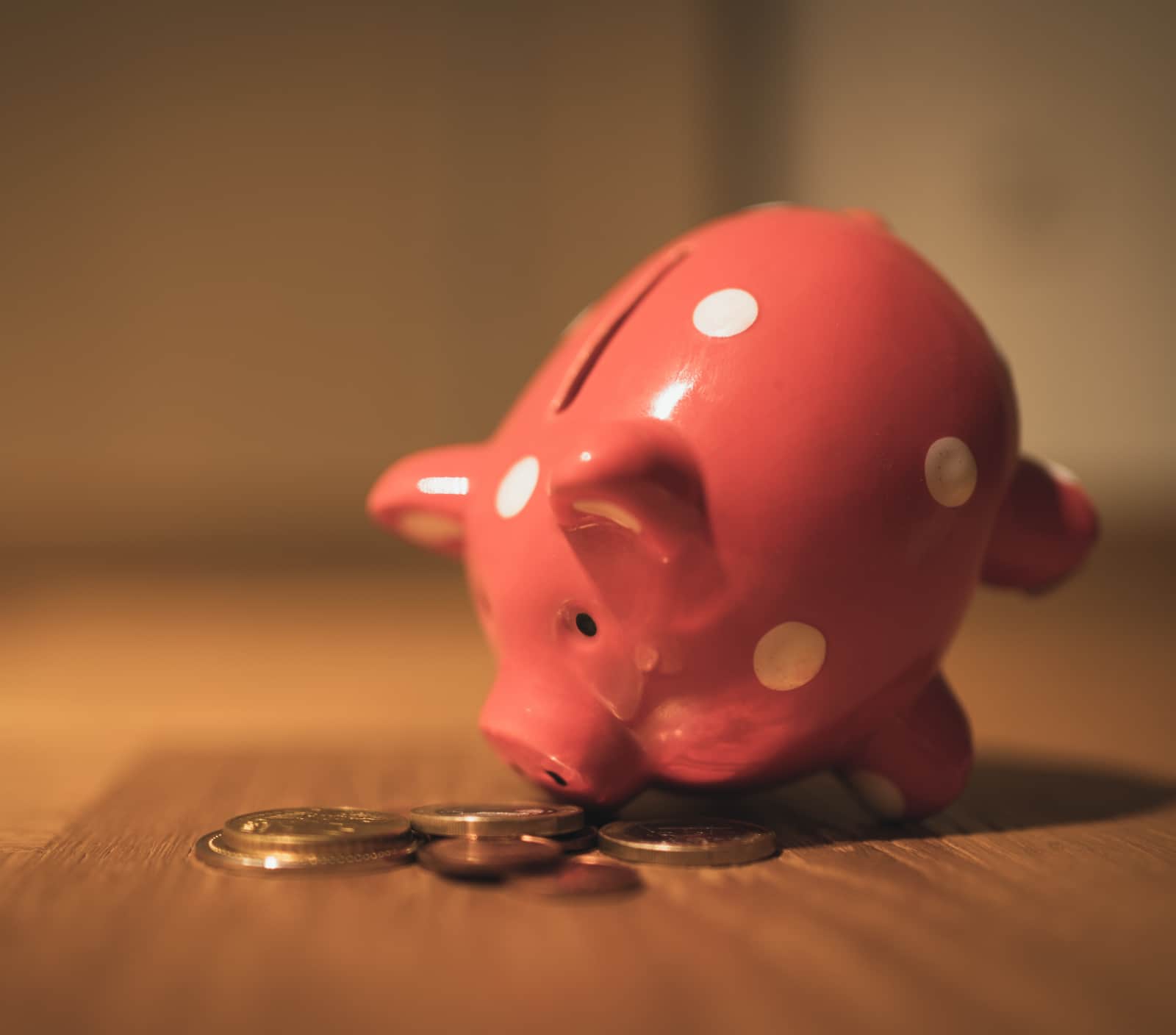 Cofrinho de porco inclinado olhando e cheirando moedas em cima de uma mesa.